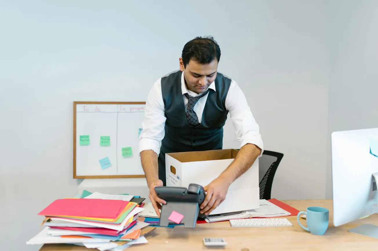Man packing office desk into a box, looking focused. Desk cluttered with colorful folders, keyboard, and mug. Whiteboard with sticky notes behind.