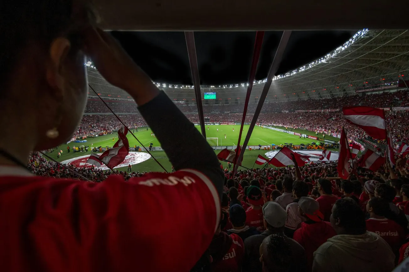 A large crowd of fans in red jerseys cheers inside a brightly lit stadium. The view shows the field and waving flags, capturing excitement and unity.