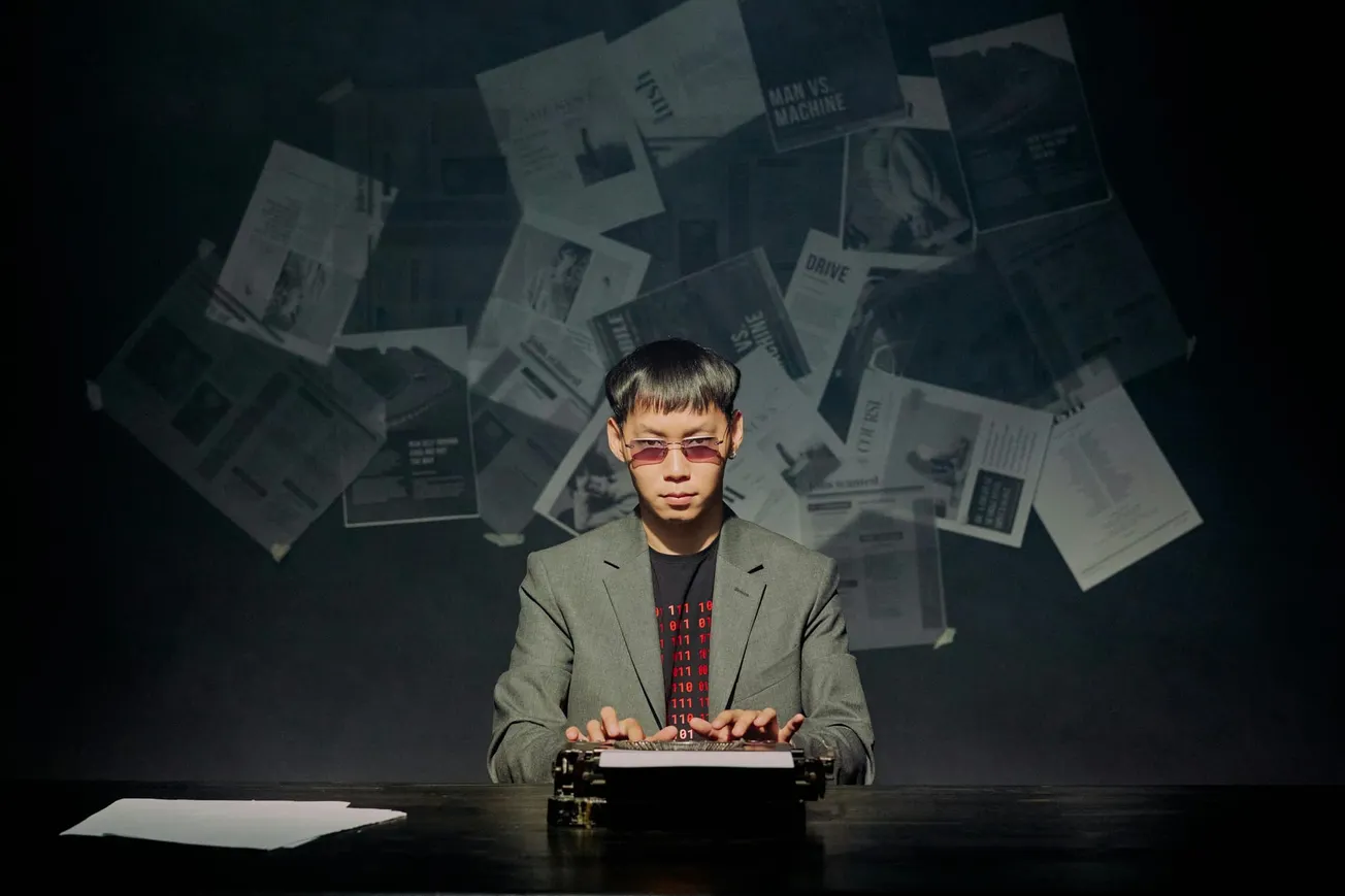 A person in a suit and sunglasses types on a typewriter against a dark background. Papers are scattered in the air, creating a mysterious, intense atmosphere.