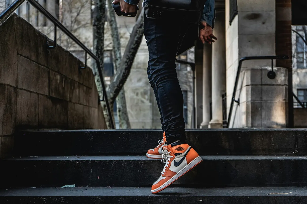 A person wearing orange and white sneakers walks up outdoor concrete steps, holding a camera. The background has stone pillars and bare trees.