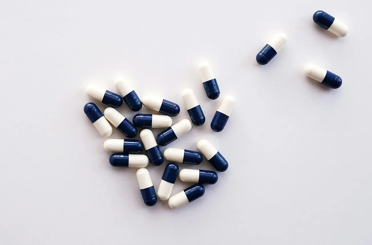 A collection of blue and white capsules scattered on a white surface, with most grouped together forming a loose heart shape, conveying a clinical tone.