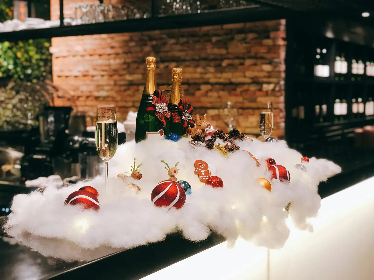 Festive bar display with champagne bottles, flutes, and Christmas ornaments on fluffy cotton resembling snow. Cozy, warm ambiance with soft lighting.