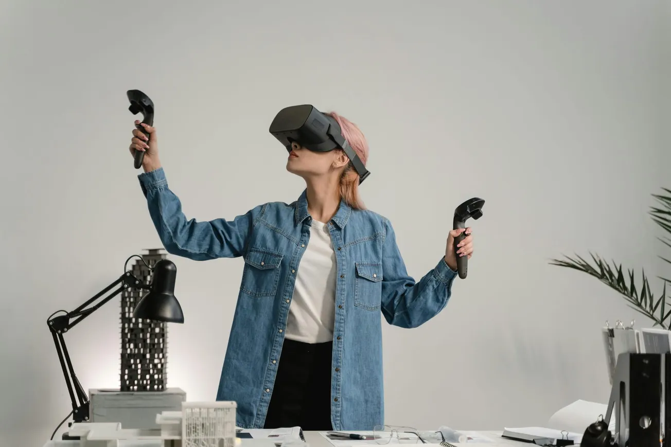 A person wearing a VR headset and denim jacket uses controllers with an immersed expression, standing by a desk with office items, conveying a tech-savvy vibe.