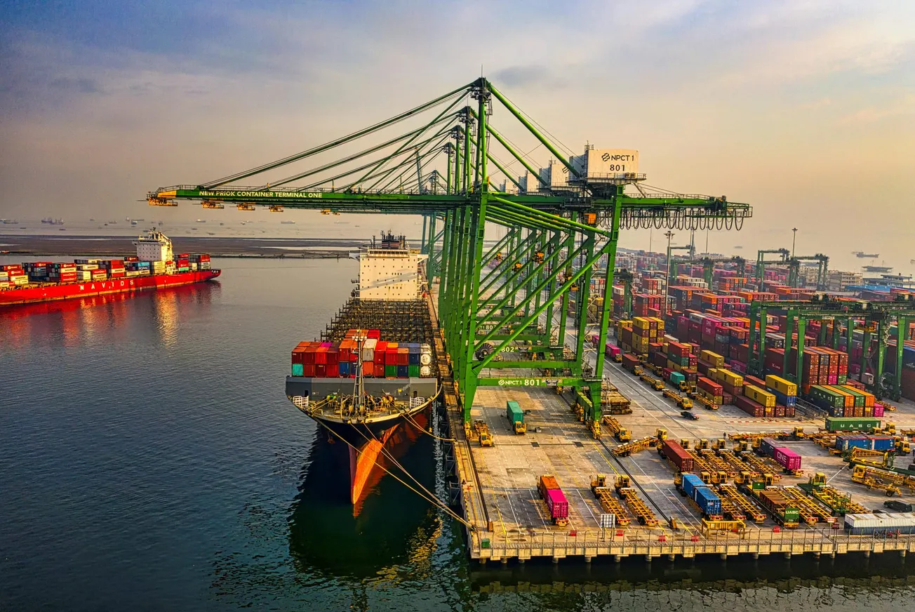 Aerial view of a bustling port with large green cranes unloading colorful containers from a docked cargo ship. Calm water surrounds, with a red ship sailing nearby.