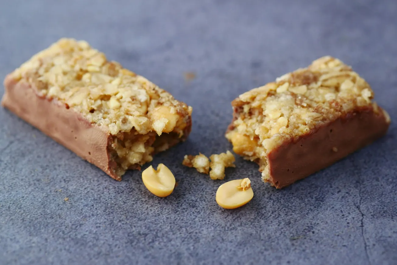 A granola bar with a chocolate-coated base is broken in half on a gray surface. Crumbs and a few peanuts are scattered around, adding texture.