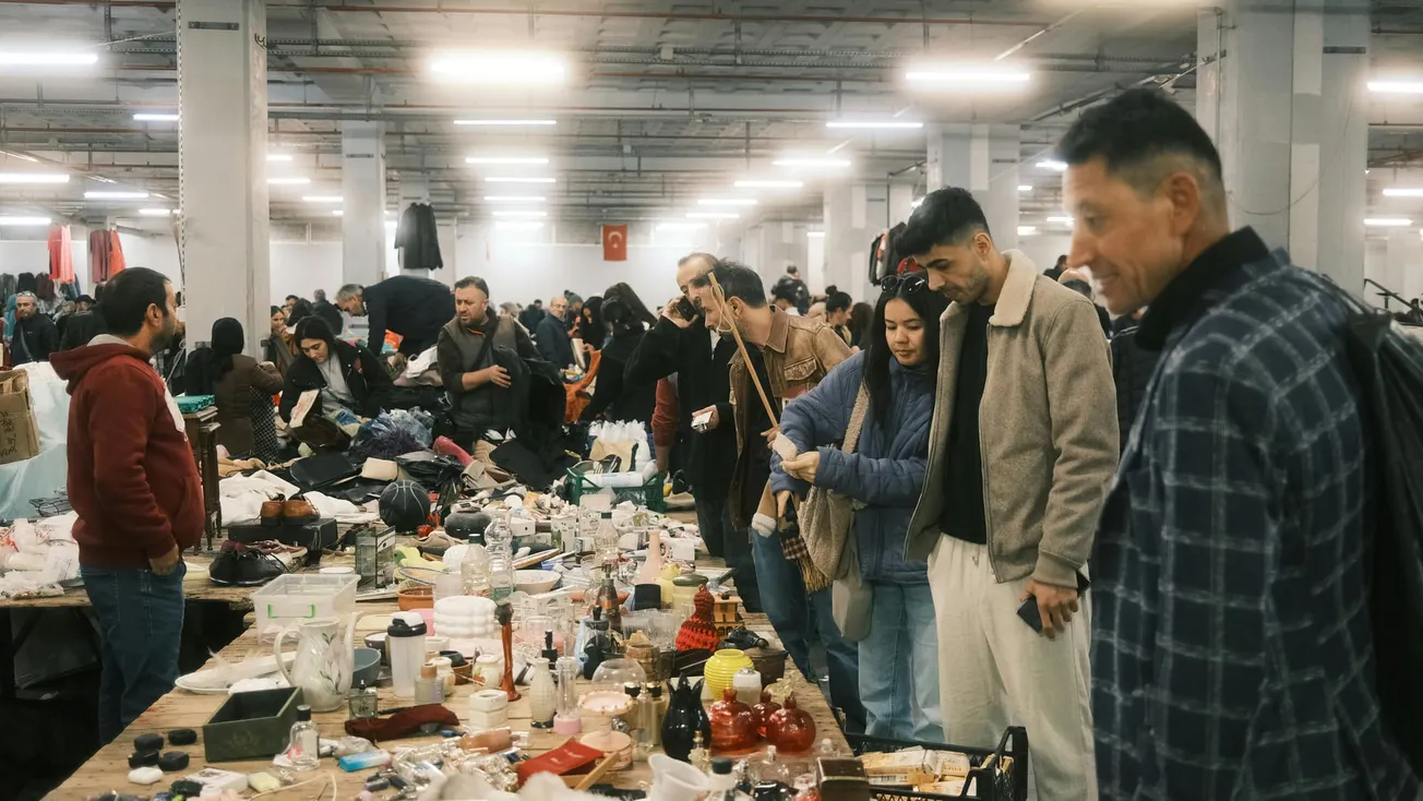 People browse goods at a busy indoor market. Tables display various items. The atmosphere is lively, with focused shoppers under bright lights.