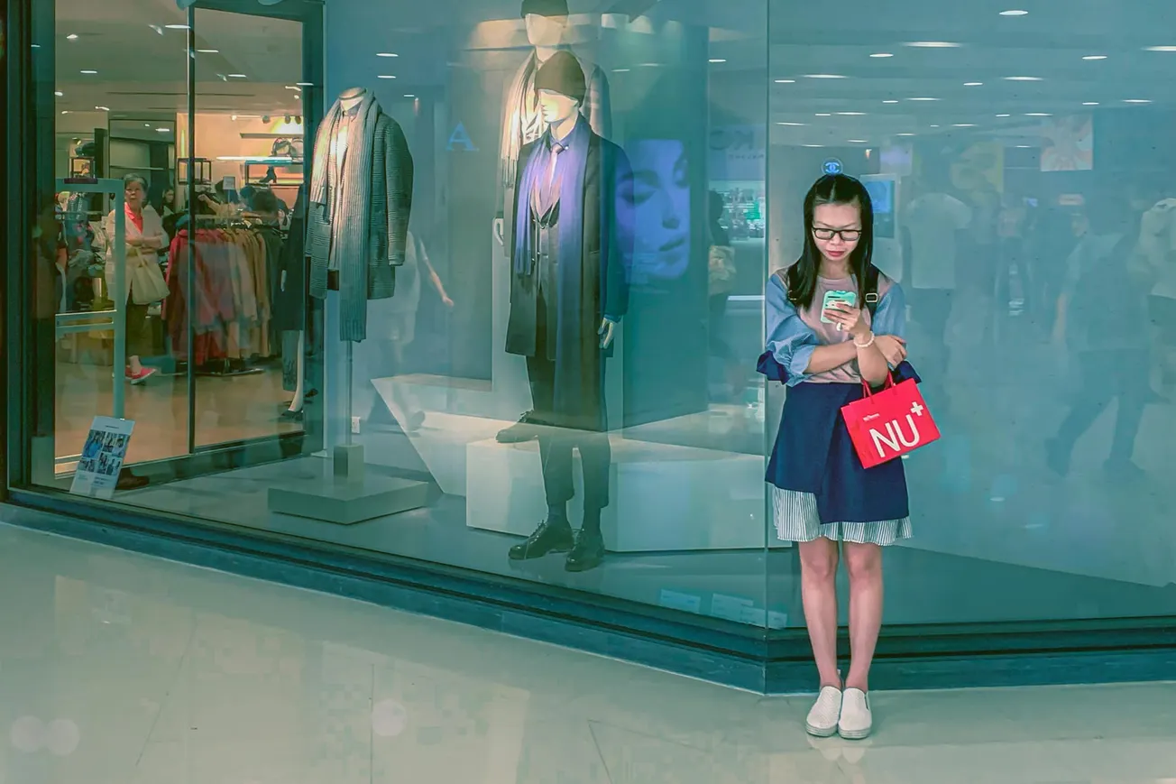 A woman stands in a mall beside a clothing store display window, engrossed in her phone. She holds a red shopping bag, with mannequins dressed in trendy attire behind her.