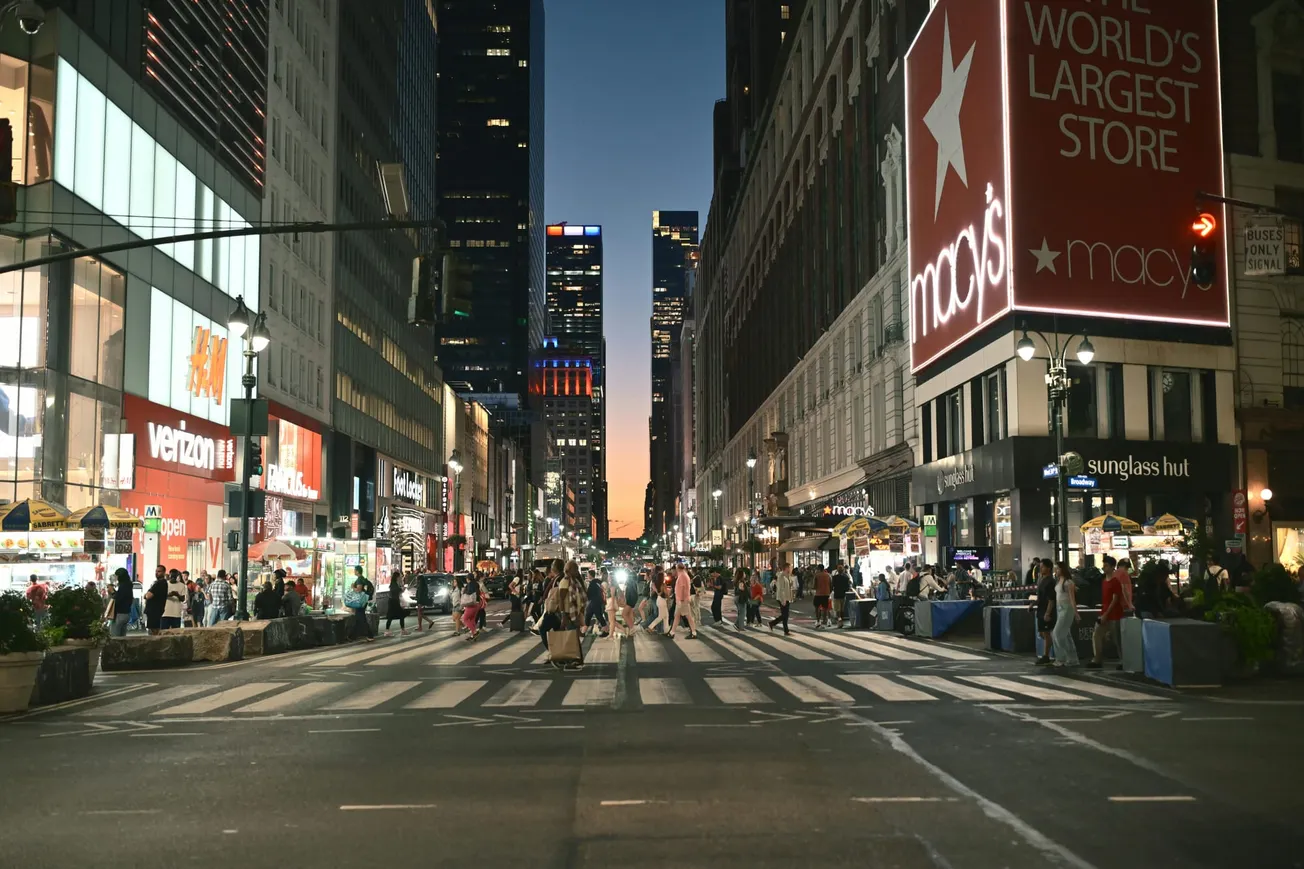Bustling city street at dusk with bright neon signs, including “Macy's.” Crowds cross the zebra-striped crosswalk, surrounded by tall buildings.