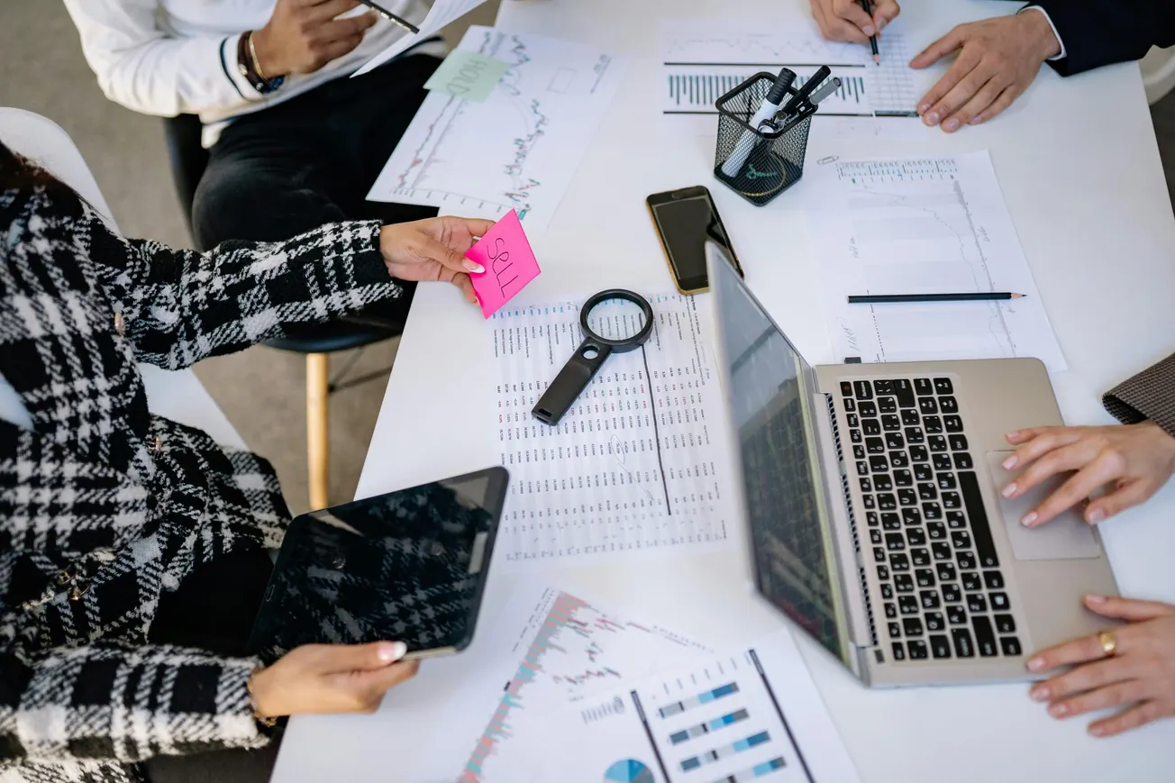 Overhead view of a collaborative office meeting. Four people around a table with charts, a laptop, magnifying glass, and notes, creating a focused, analytical atmosphere.