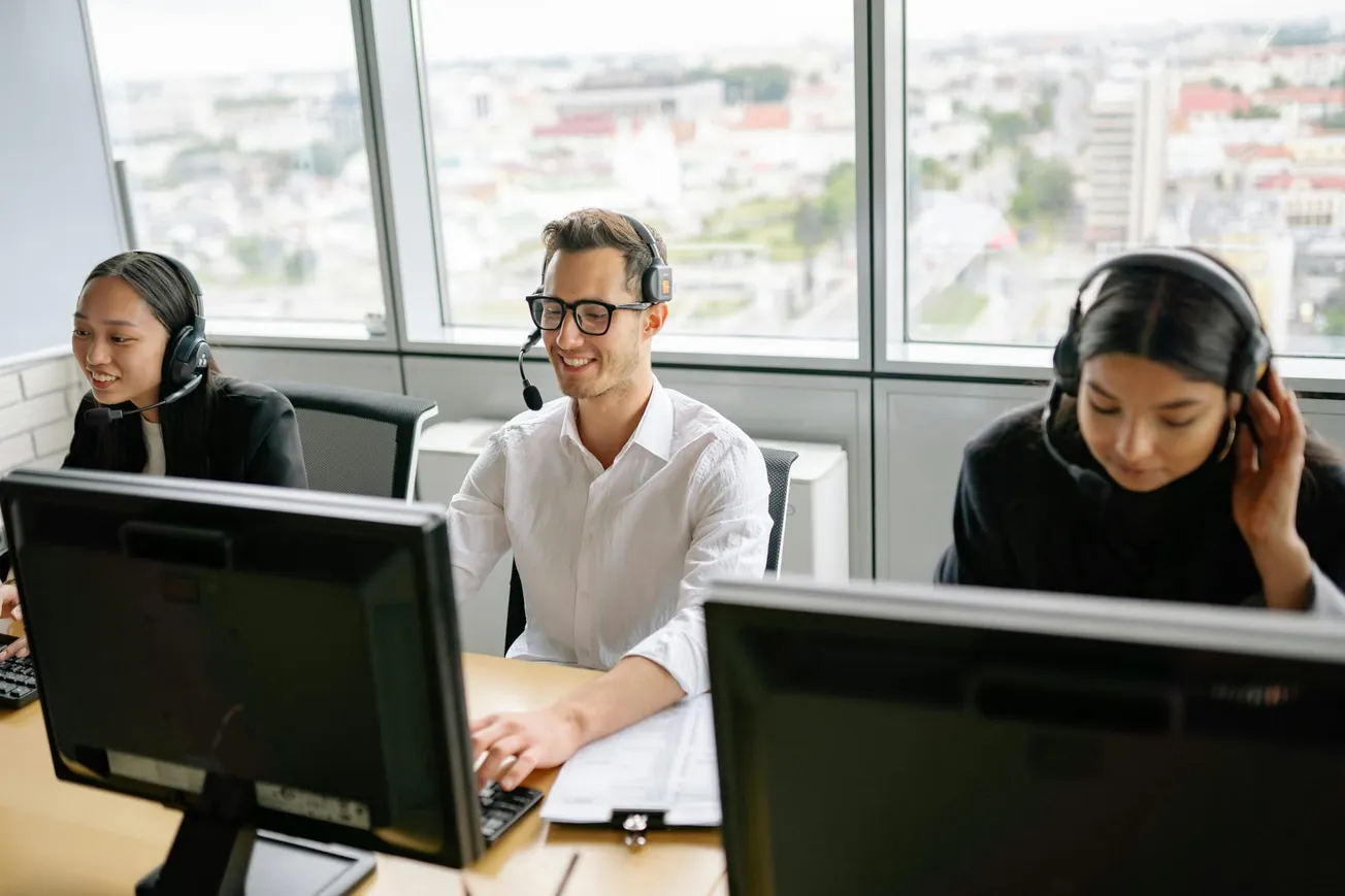 Three call center operators wearing headsets work at computers in a bright office with large windows. They seem focused and engaged.