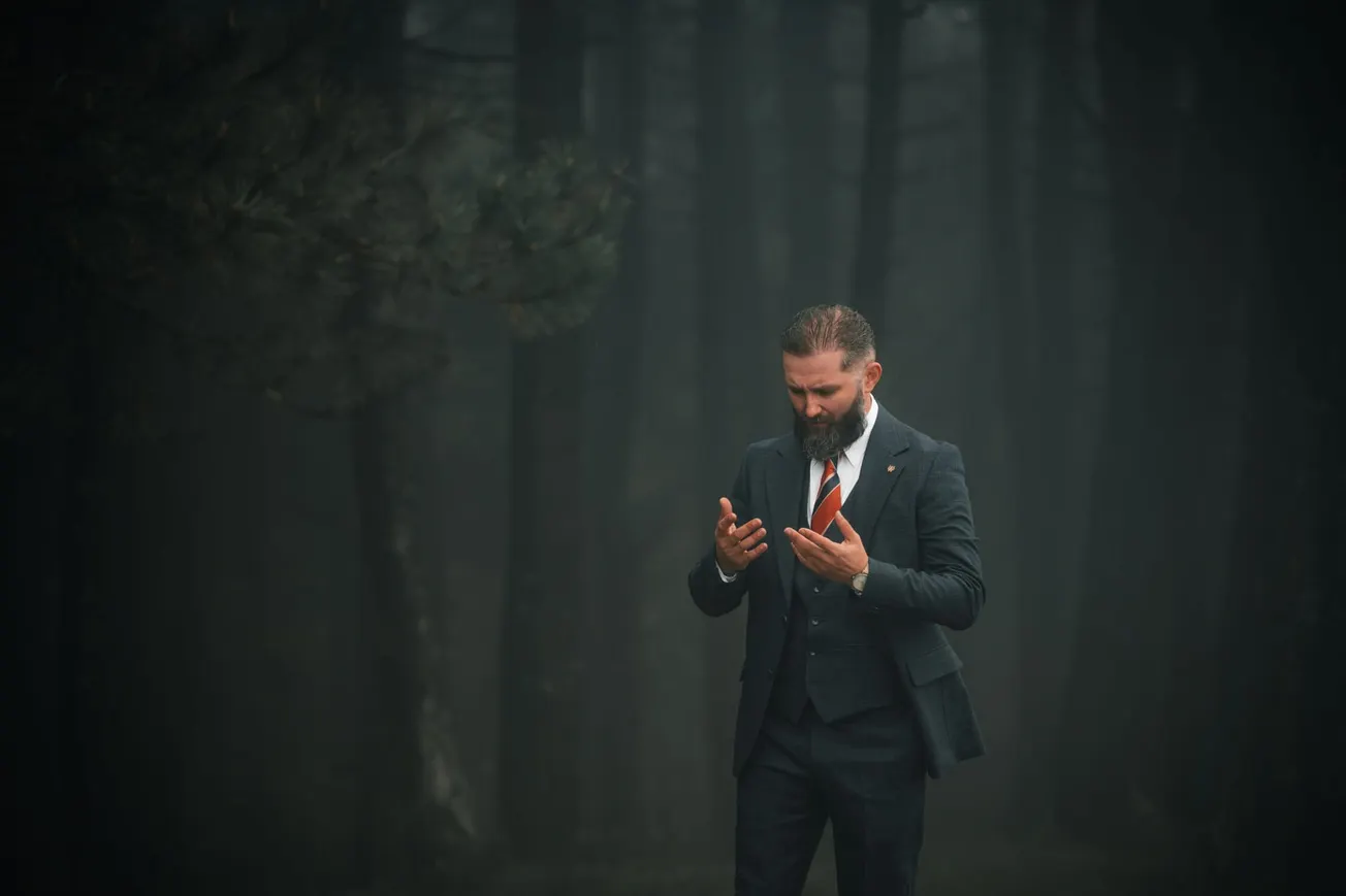 A bearded man in a dark suit with a red tie stands in a misty forest, looking contemplative with his hands raised. The mood is mysterious and introspective.