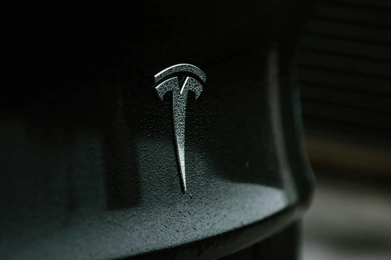 Close-up of a silver Tesla logo on a sleek, dark surface. The reflective material and soft lighting convey a modern and sophisticated feel.