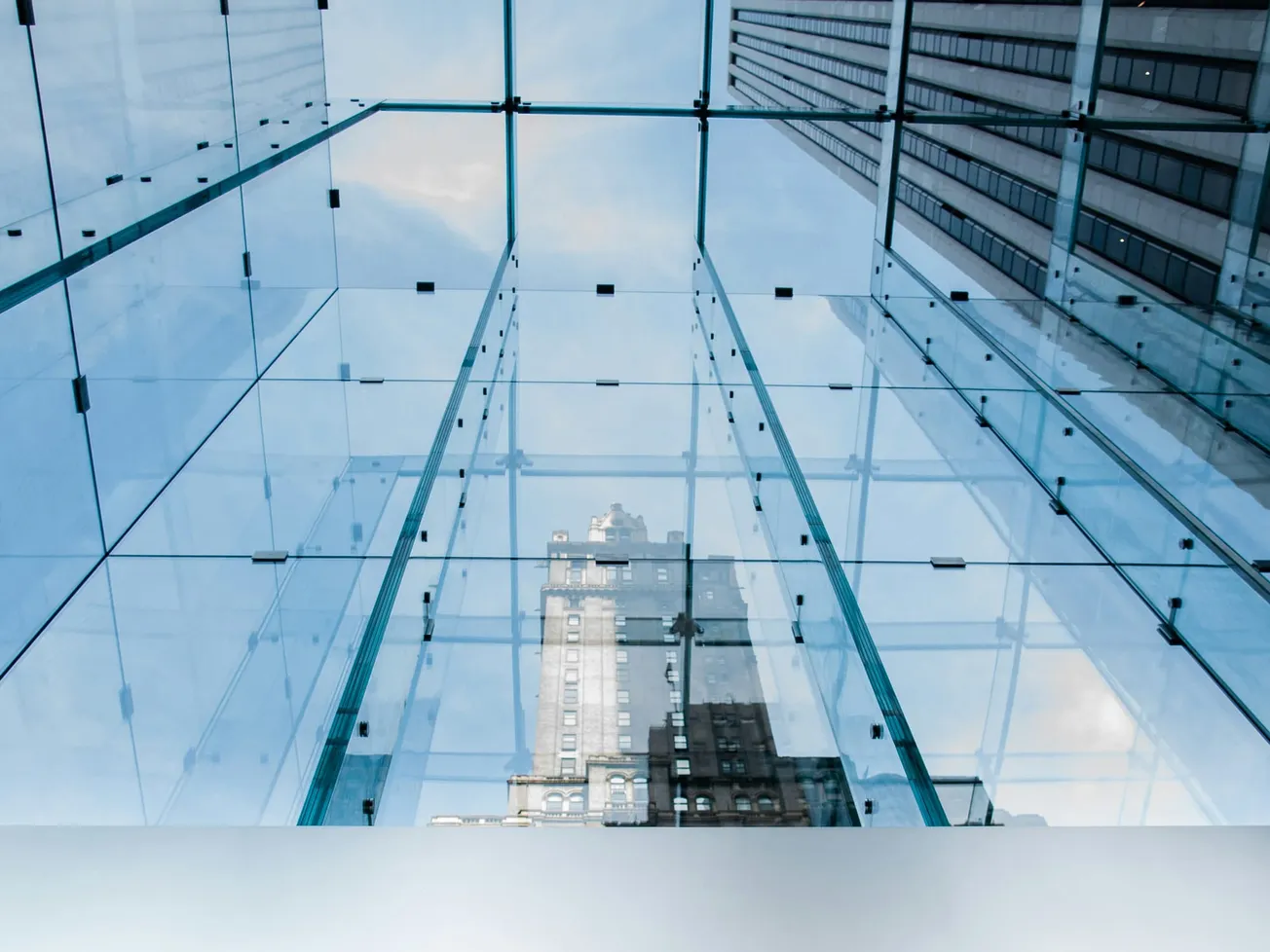 View of a city skyscraper through a reflective glass ceiling and walls with blue sky, evoking modernity and urban sophistication.