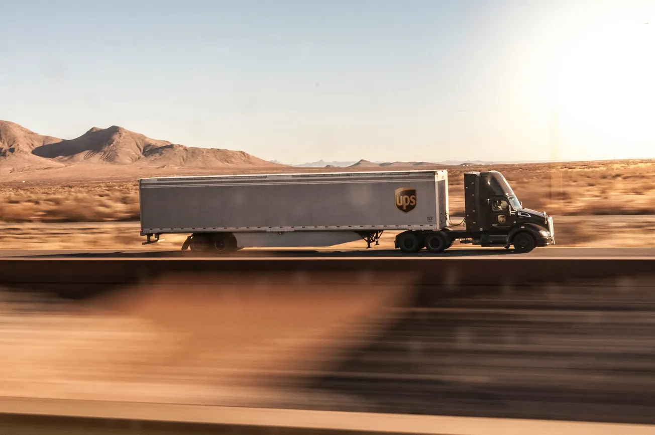A UPS semi-truck drives along a highway through a sunlit desert landscape. Mountains are visible in the background, conveying a sense of vastness and speed.