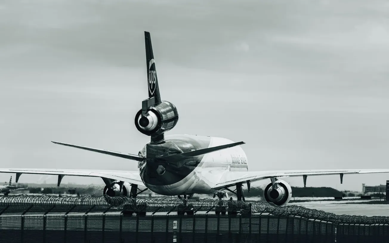 Monochrome image of a parked cargo aircraft, viewed from the rear, on an airport runway. Overcast sky above, with a distant landscape visible. Calm atmosphere.