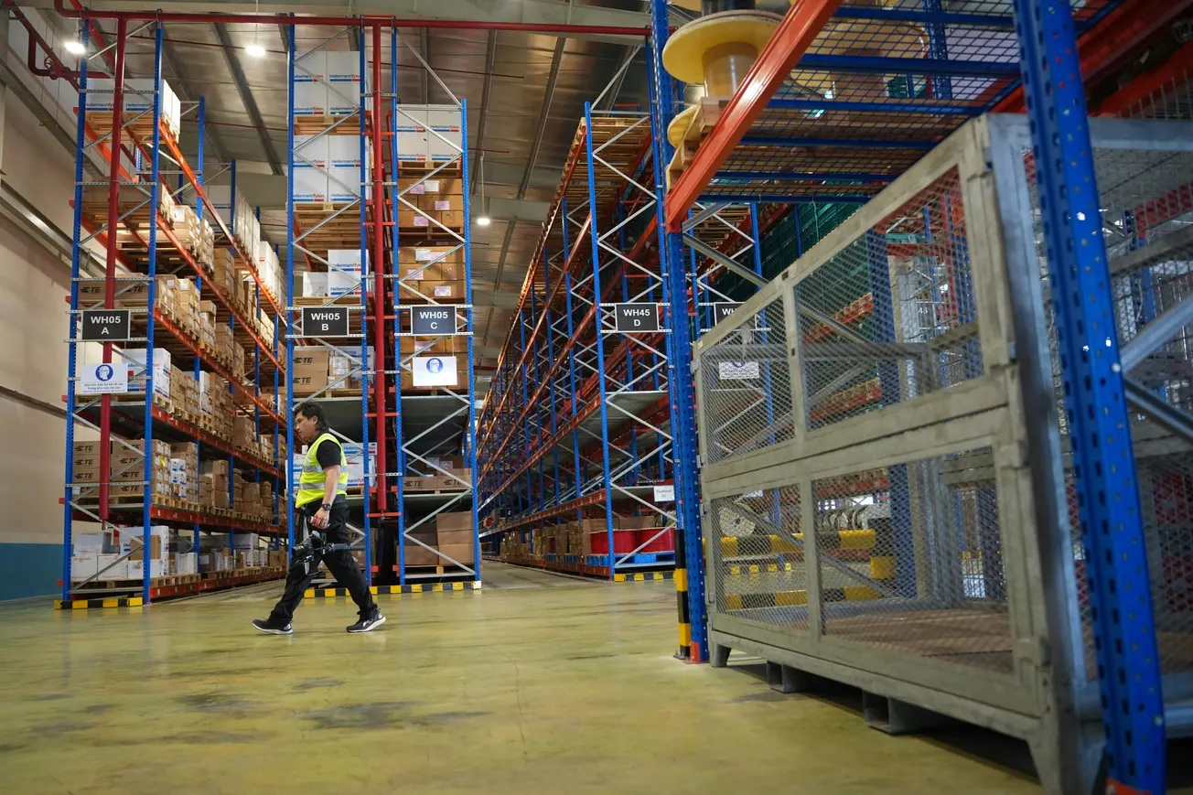 A worker in a yellow safety vest walks through a spacious warehouse with tall blue and orange shelves full of neatly stacked boxes, conveying efficiency.