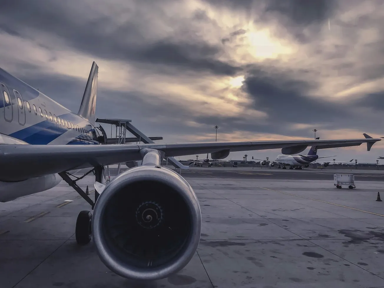 Sunset over an airport with a close-up of a parked airplane's wing and engine in the foreground. The sky is dramatic with clouds, conveying a calm mood.