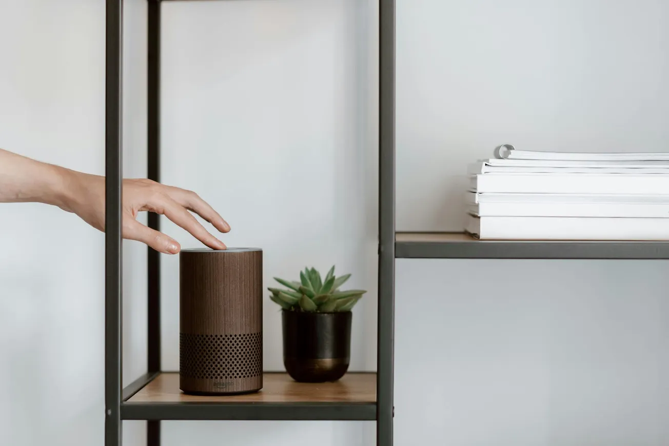 A hand reaches for a smart speaker on a minimalist wooden shelf, beside a small green succulent. The setting is calm and modern, with white walls.