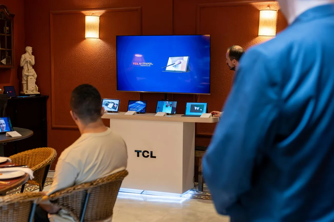 In a warmly lit room, people watch a presentation featuring TCL's latest technology on a large screen. Various devices are displayed on a stand.