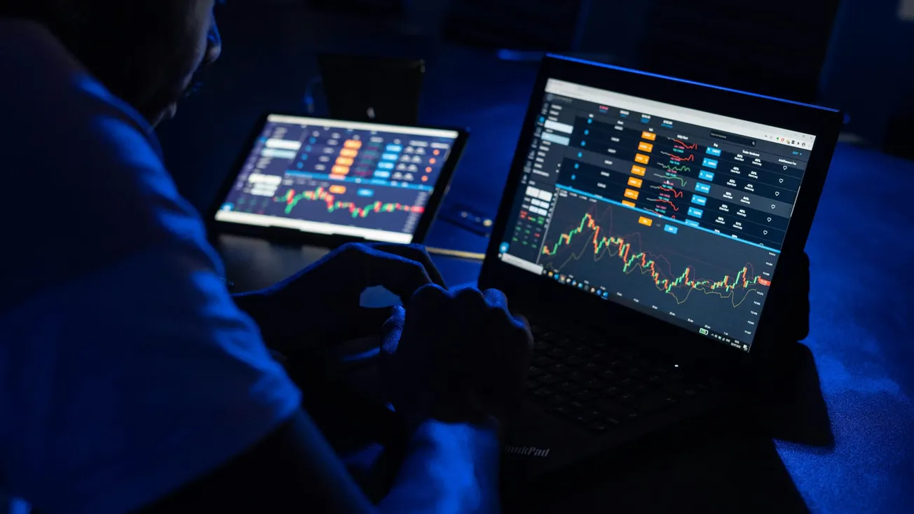 A person in dim lighting views two screens displaying financial graphs and stock market data, conveying a focused and analytical mood.