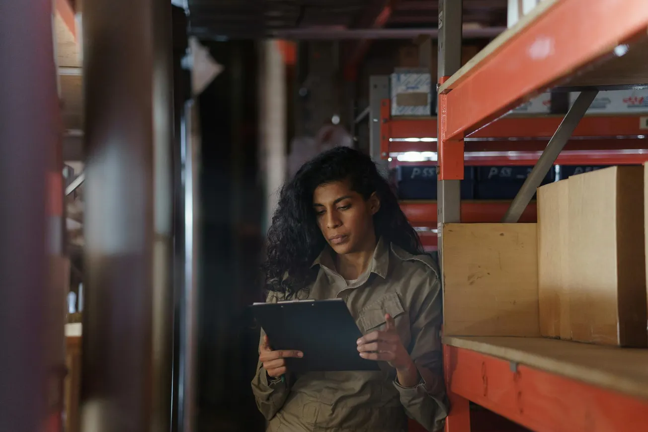 Woman in a warehouse checks inventory on a clipboard. She stands next to shelves and boxes, focused and thoughtful, in a dimly lit space.