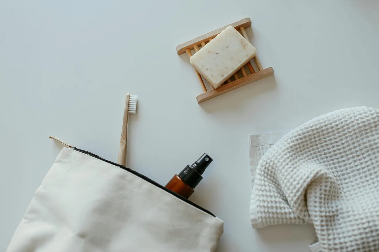 Top view of eco-friendly bathroom items on a white surface, including a bamboo toothbrush, soap on a wooden tray, spray bottle, and beige waffle towel.