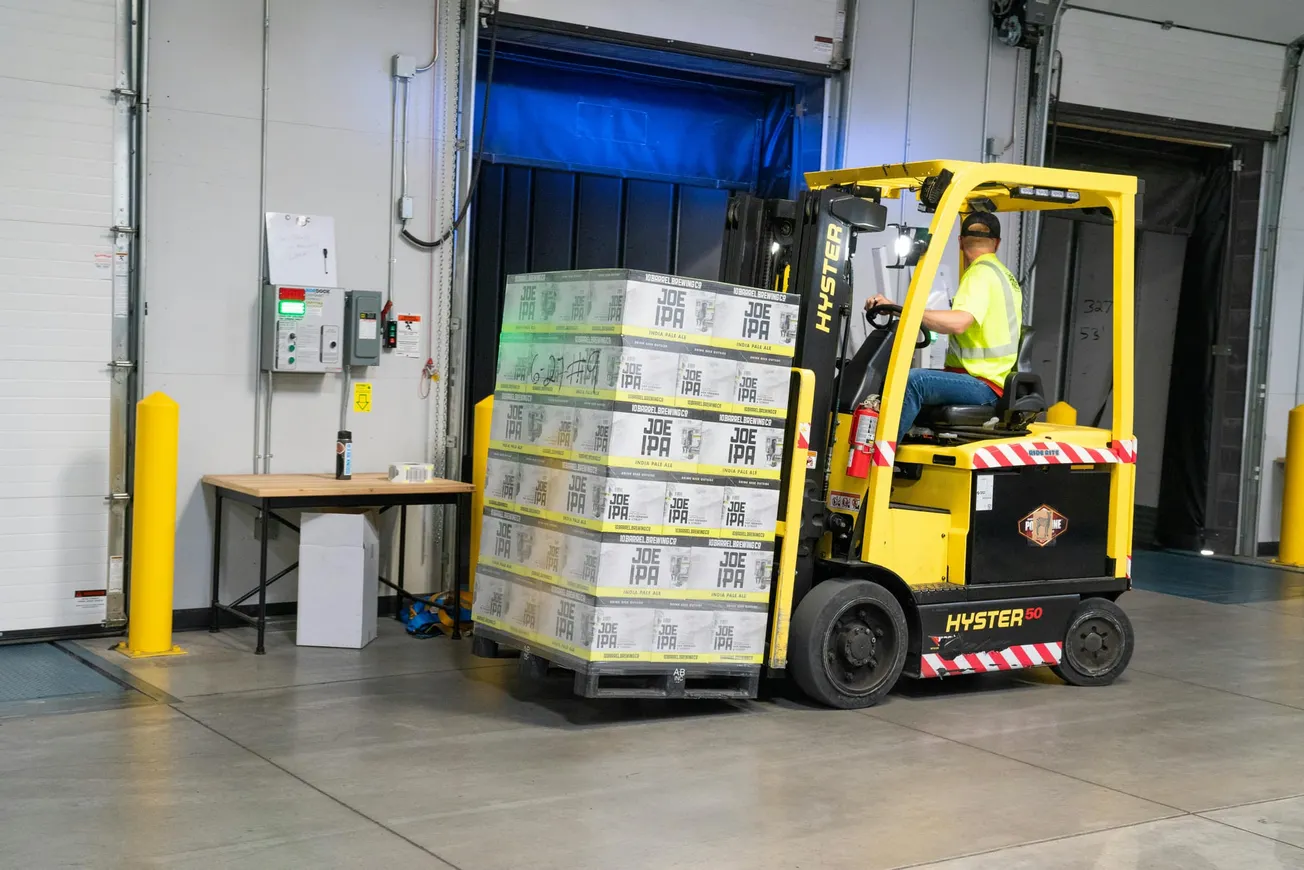 A worker operates a yellow forklift, unloading a pallet of boxed "Joe IPA" beer in a warehouse loading dock. A safety vest and caution markings are visible.