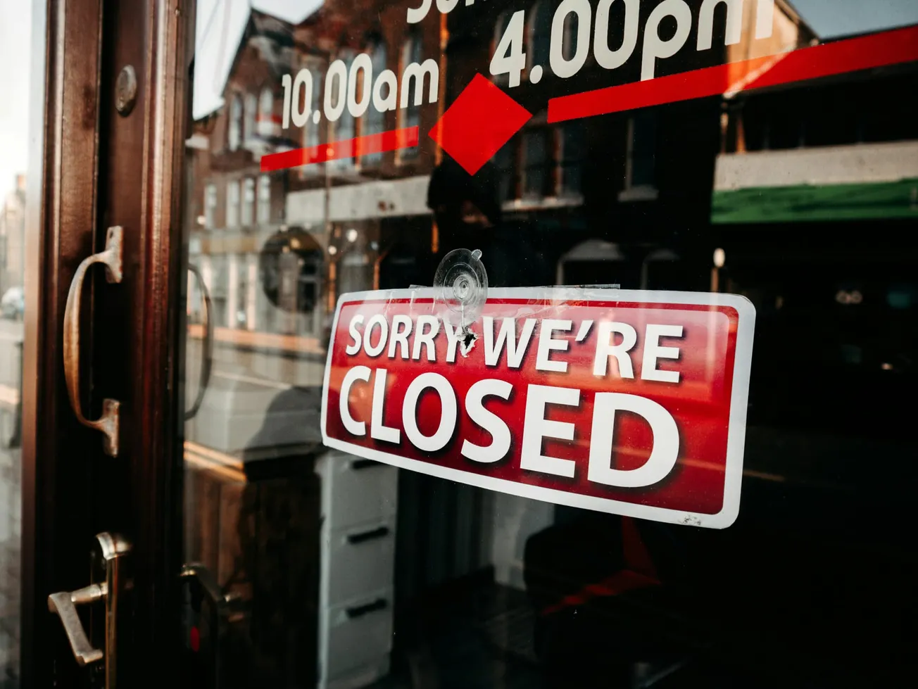 Storefront glass door with a red "Sorry, We're Closed" sign. Reflections show buildings and a street.