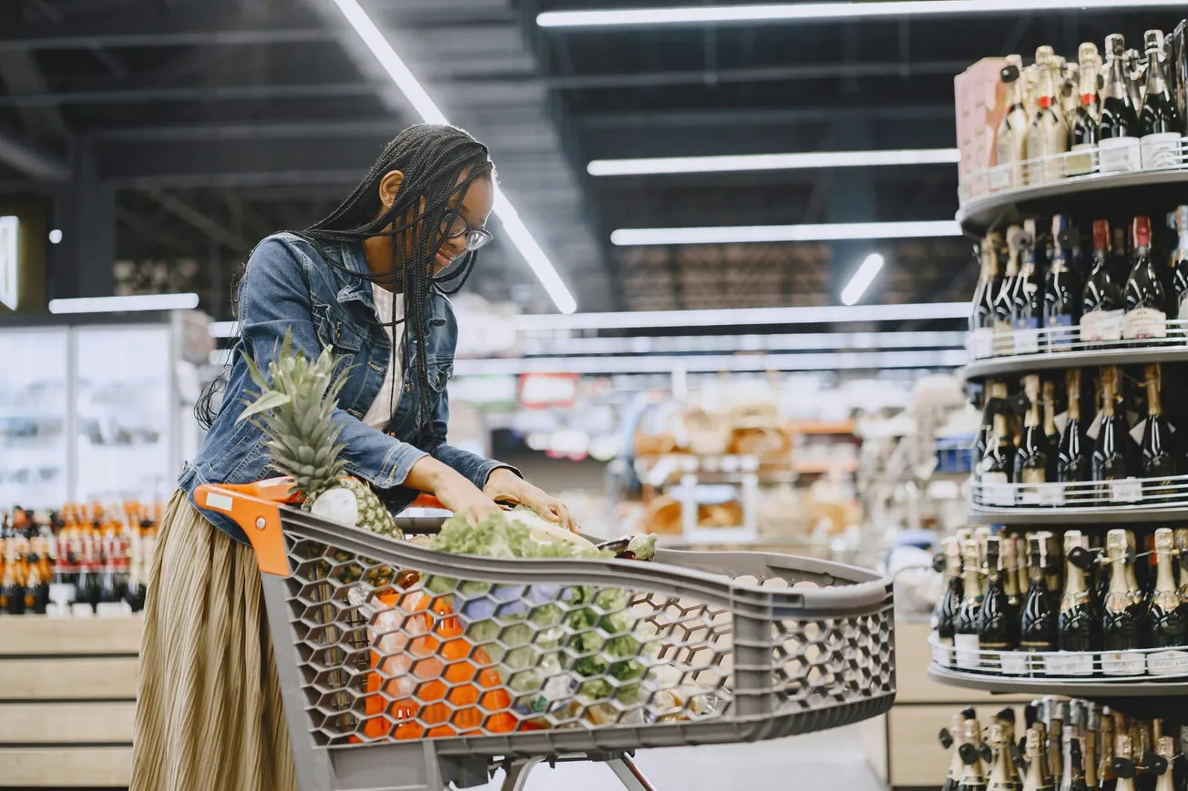 A woman in a denim jacket shops in a supermarket, pushing a cart full of produce, including pineapples and oranges, with wine bottles on shelves nearby.