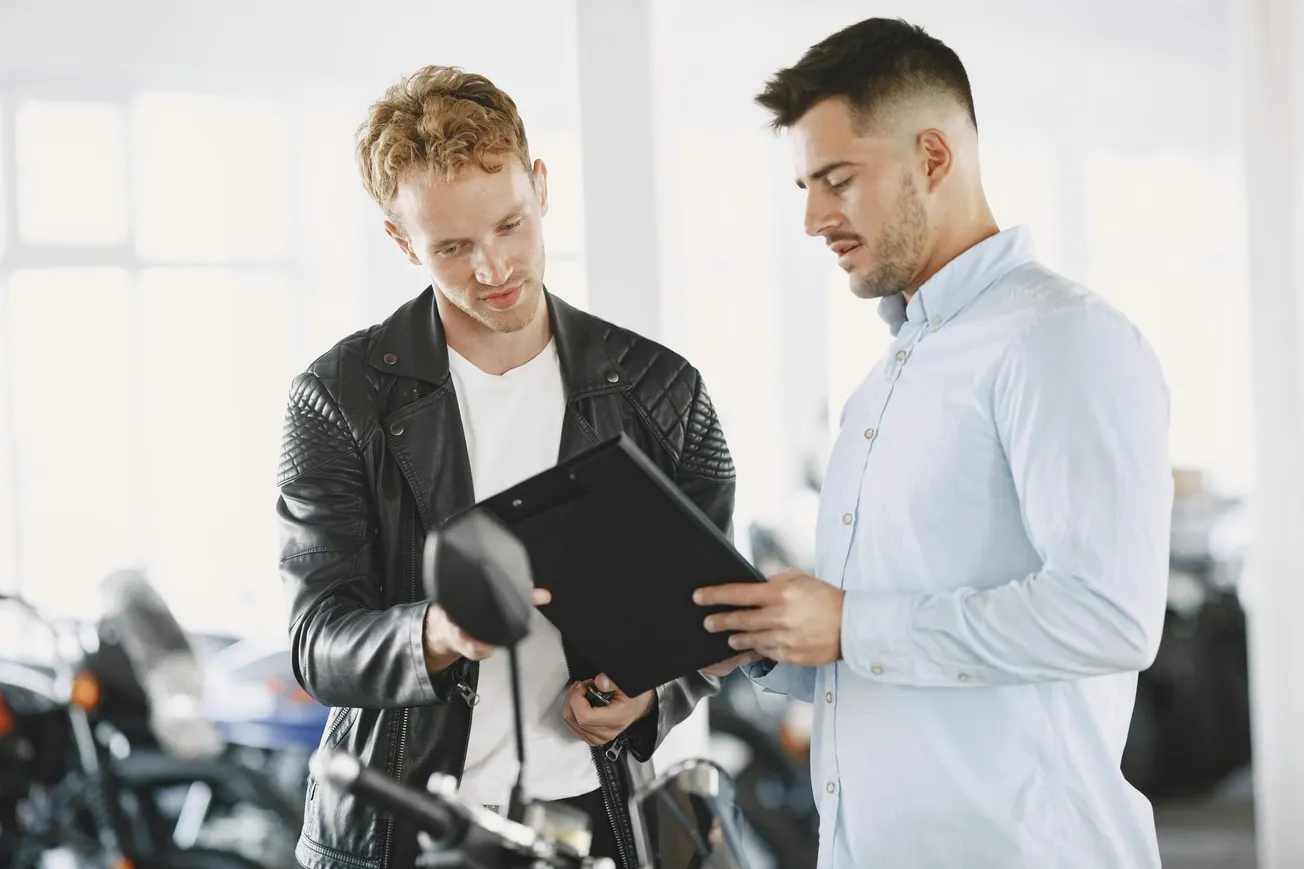 Two men stand in a motorcycle showroom. One in a leather jacket examines a clipboard held by the other in a light blue shirt, discussing details.