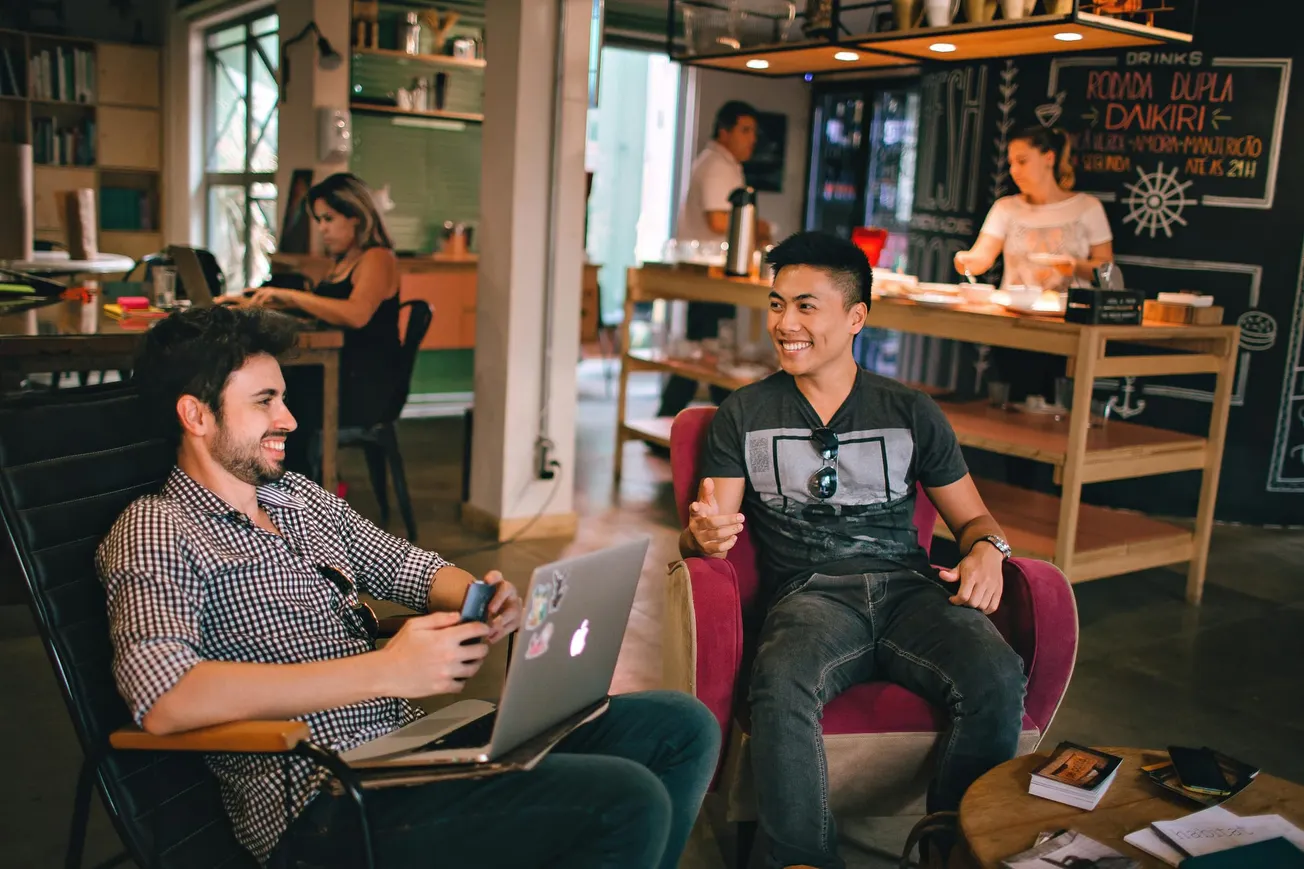 Two men engage in lively conversation at a café, each seated with a laptop. In the background, two people work at a counter, creating a relaxed and social atmosphere.