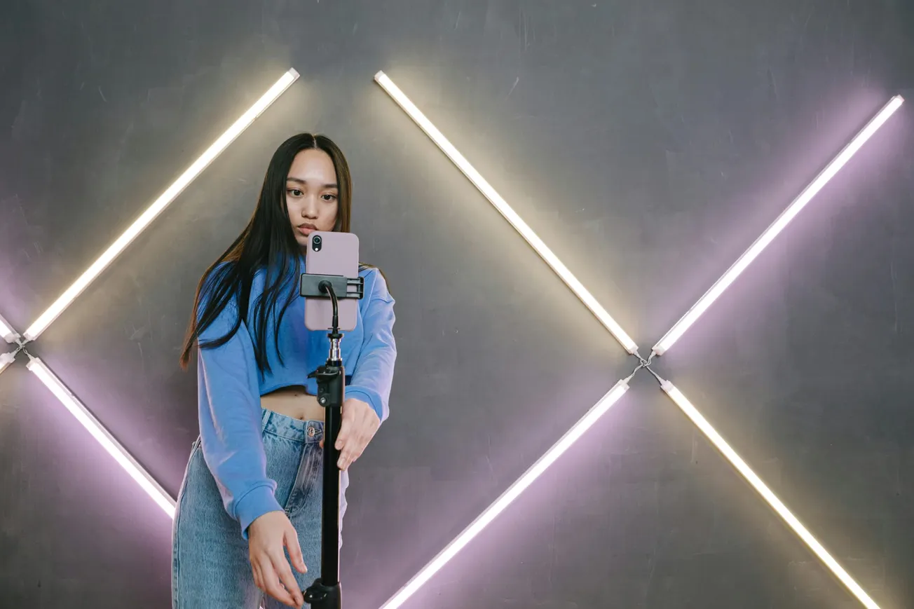 A woman in a blue sweater and jeans takes a selfie with a smartphone on a tripod. She stands before a wall with glowing X-shaped neon lights, creating a modern and vibrant atmosphere.