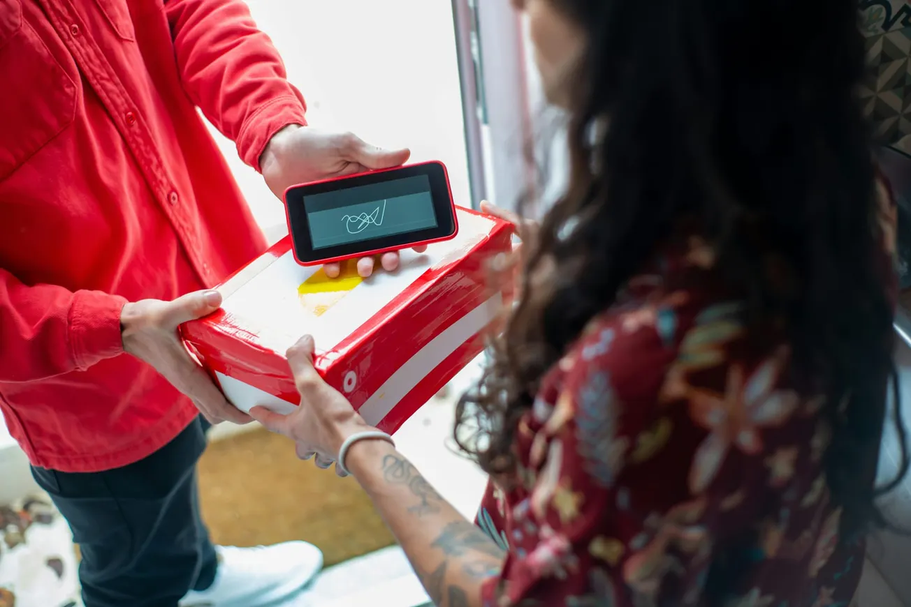A delivery person in a red jacket holds a package and a digital signature pad. A woman, wearing a floral shirt, signs for the package at the door.