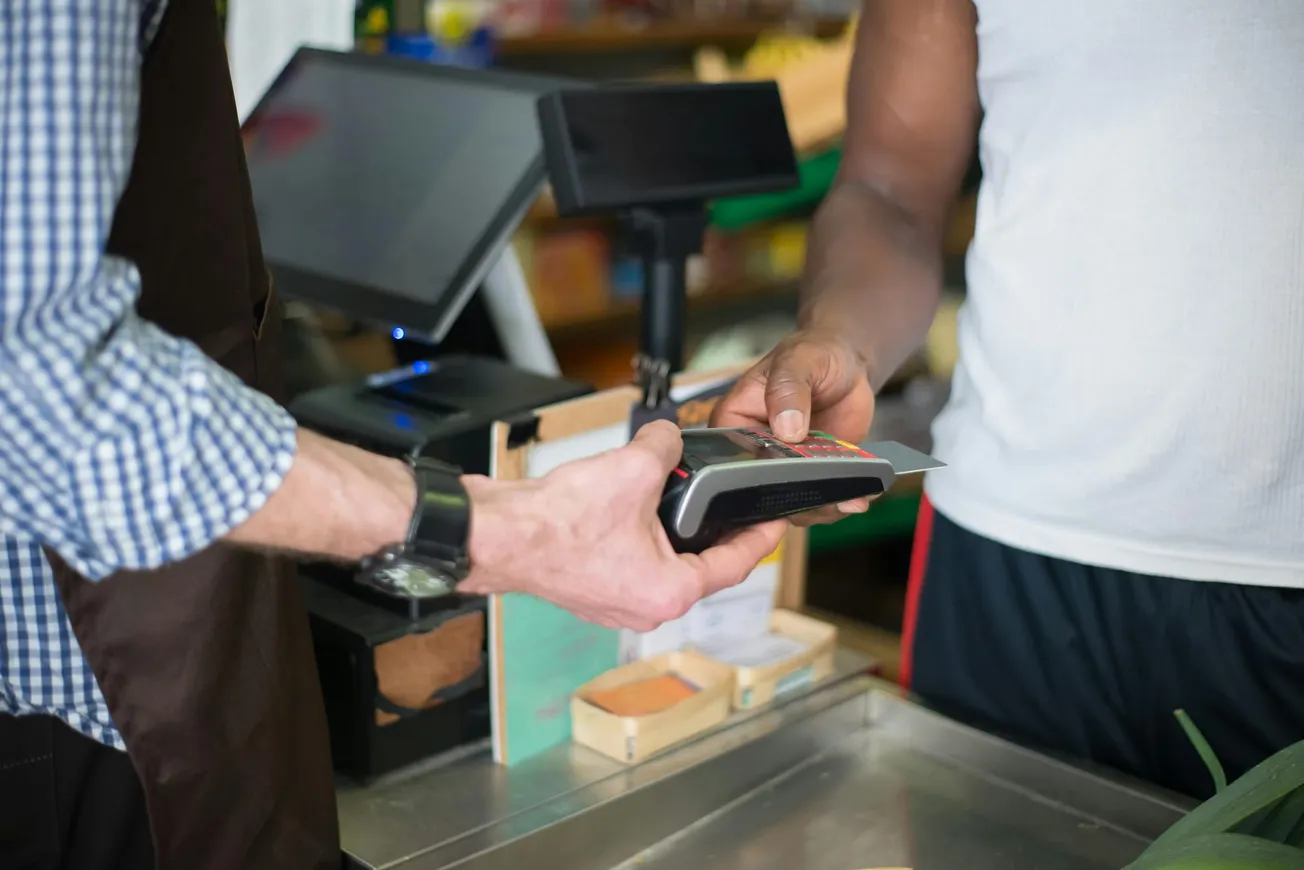 A customer in a white shirt uses a credit card for contactless payment at a checkout counter, where a cashier in an apron holds the terminal.