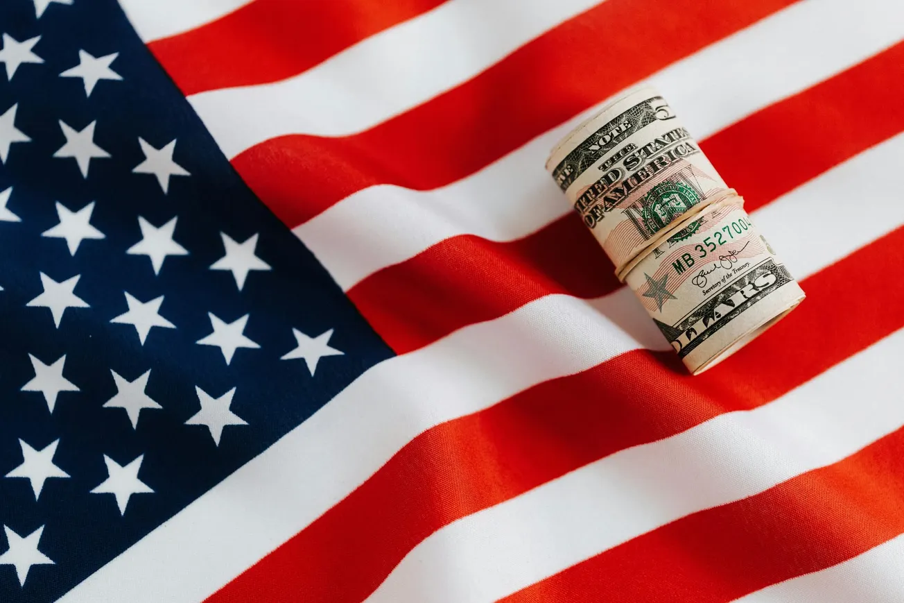 A rolled-up stack of U.S. dollar bills sits on a waving American flag. The image conveys themes of finance, patriotism, and economic pride.