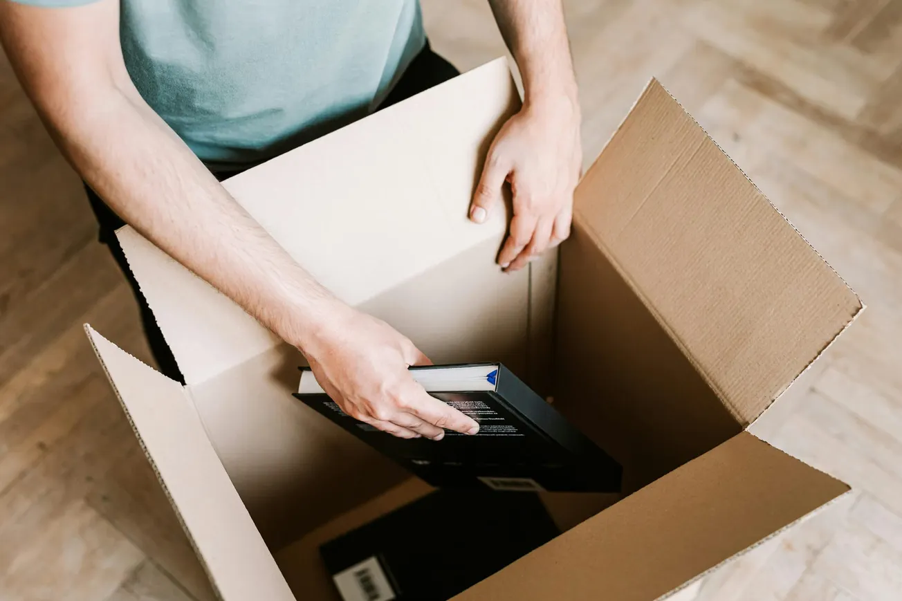 A person places a book inside an open cardboard box on a wooden floor. The scene suggests packing or organizing, with a calm, focused mood.