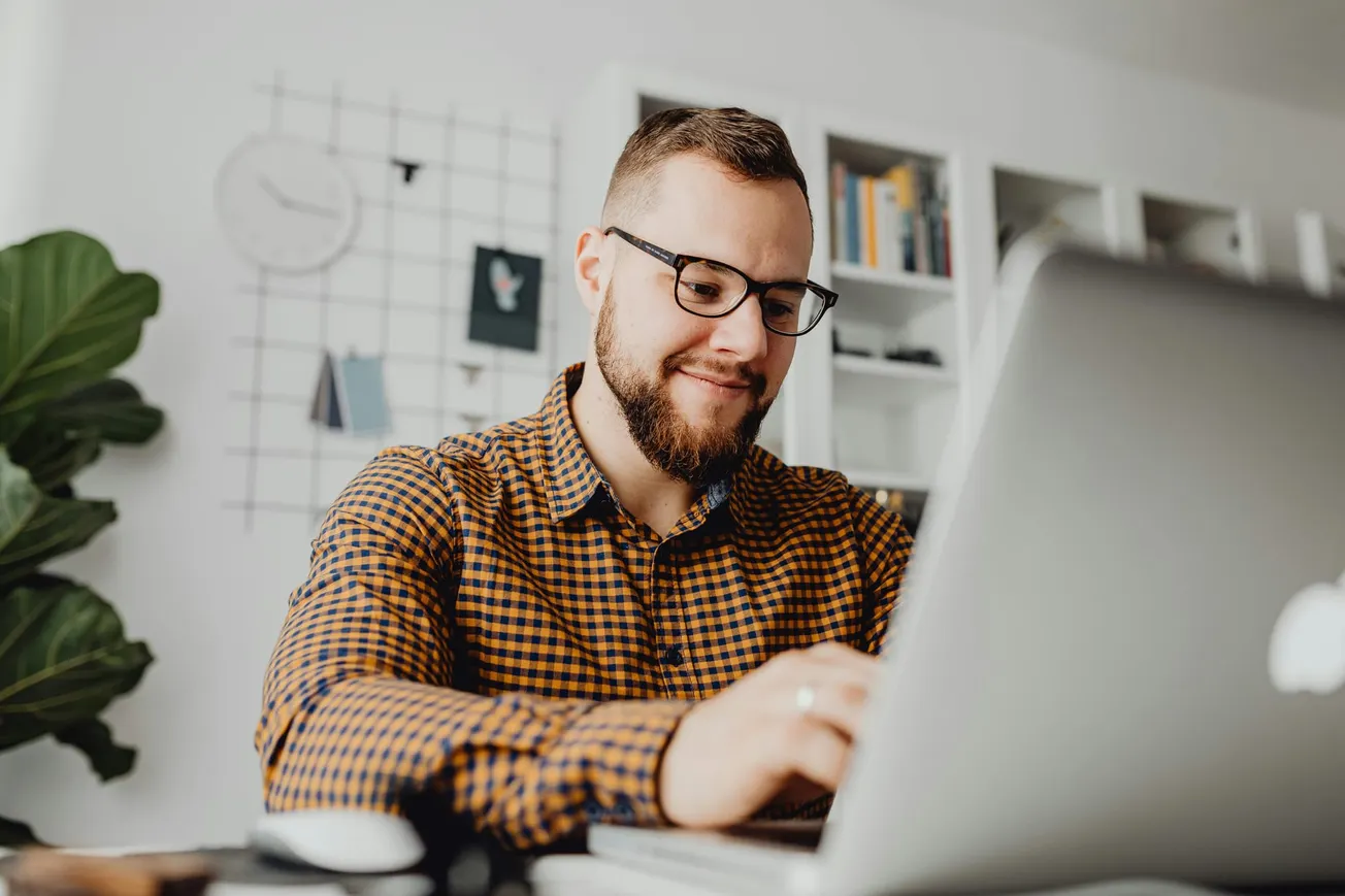 A man with glasses and a beard smiling while typing on a laptop, sits at a desk in a home office. Bookshelves and a large plant are in the background.