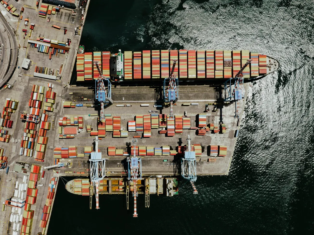 Aerial view of a bustling cargo port with colorful shipping containers and cranes on a dock. Sunlit water surrounds the area, creating a dynamic, busy atmosphere.