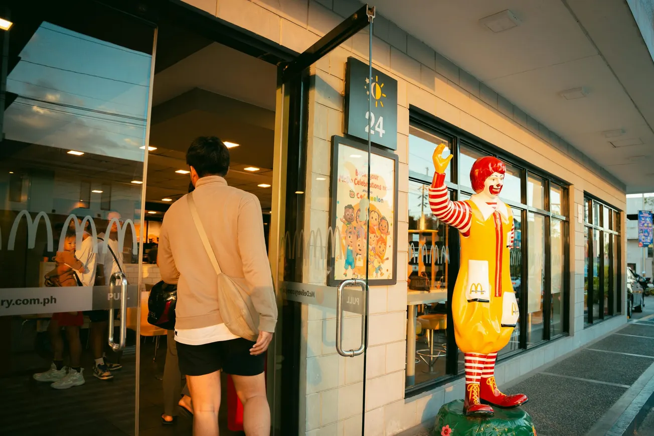 A person enters a brightly lit fast food restaurant. A Ronald McDonald statue waves outside. The scene conveys a welcoming, casual atmosphere.