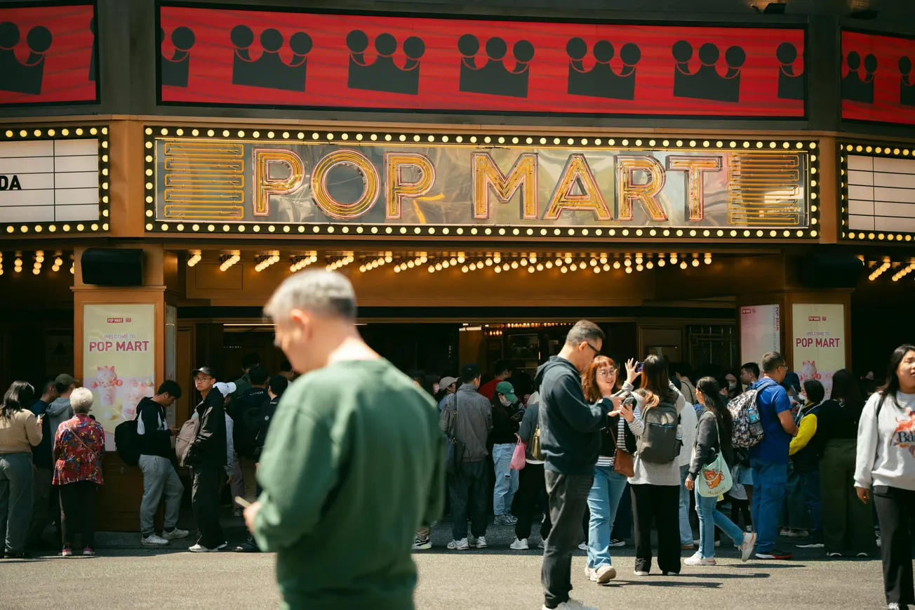 A crowd gathers outside a brightly lit theater with a marquee sign reading "Pop Mart." People chat and look at their phones, creating a lively, bustling atmosphere.