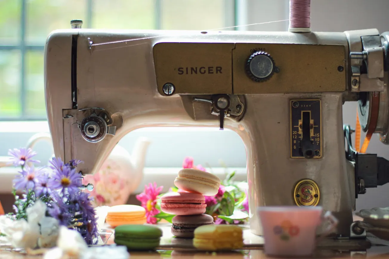 A vintage sewing machine set against a window, surrounded by pastel macarons, purple flowers, and a teacup, creating a nostalgic, cozy atmosphere.