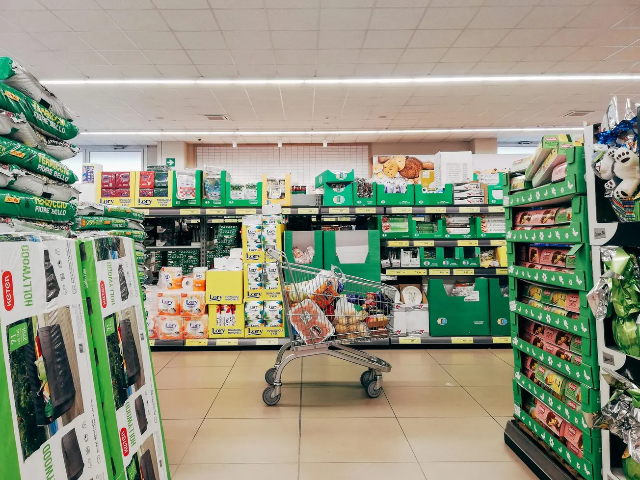 A shopping cart filled with groceries is in the aisle of a brightly lit supermarket. Shelves are stocked with assorted packaged goods and household items.
