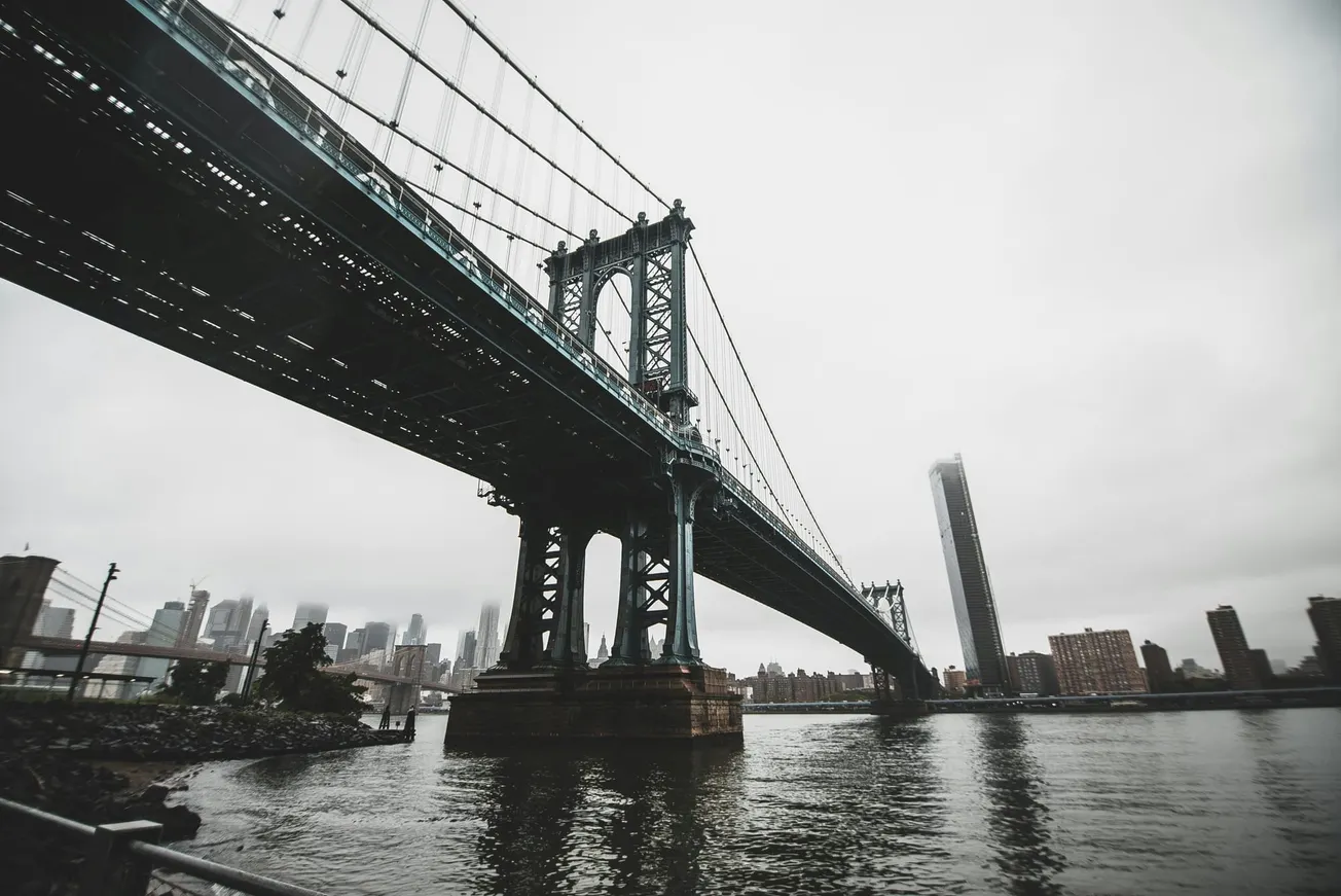 Suspension bridge spanning a river under a cloudy sky, with a city skyline in the background. The scene conveys a moody and majestic atmosphere.
