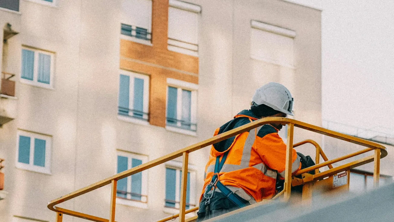 A construction worker in a bright orange safety jacket and helmet operates a lift, against the backdrop of a multi-story building with balconies.