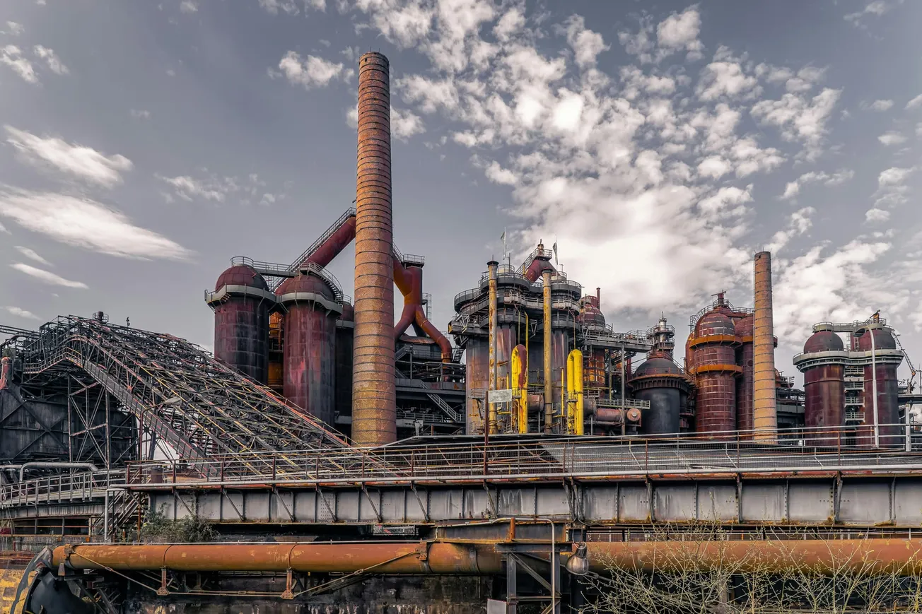 Alt text: A large, rusted industrial complex with towering chimneys and pipes set against a partly cloudy sky. The scene conveys an abandoned, historical atmosphere.