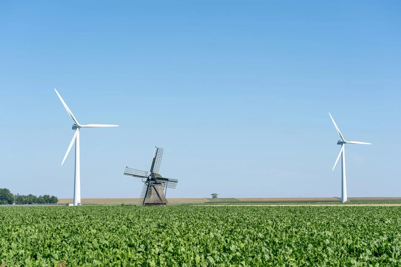 Two modern wind turbines and a traditional windmill in a green field under a clear blue sky, symbolizing the contrast between old and new energy sources.