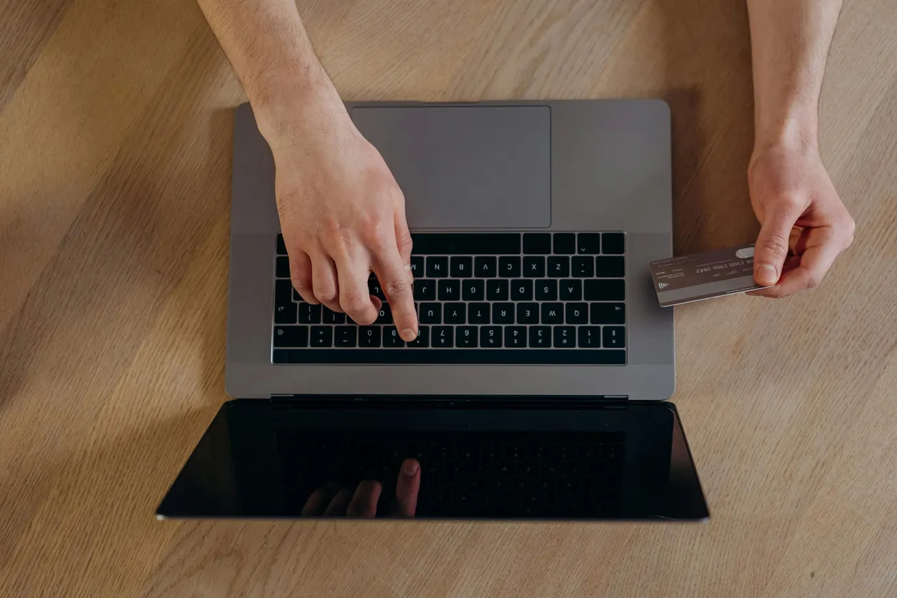 A person is using a laptop on a wooden table, holding a credit card in one hand and typing on the keyboard with the other, suggesting online shopping.