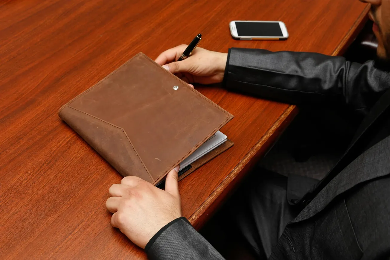 A person in a suit holds a pen and a brown leather folder at a wooden table, with a smartphone nearby, conveying a professional setting.