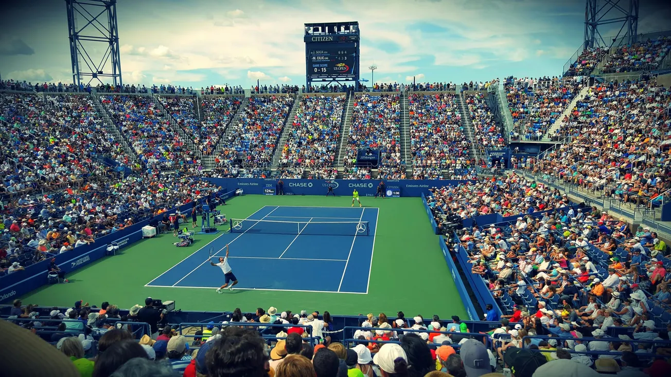 A packed tennis stadium with a vibrant crowd surrounds a match in progress on a bright day. A player prepares to serve on the blue and green court.