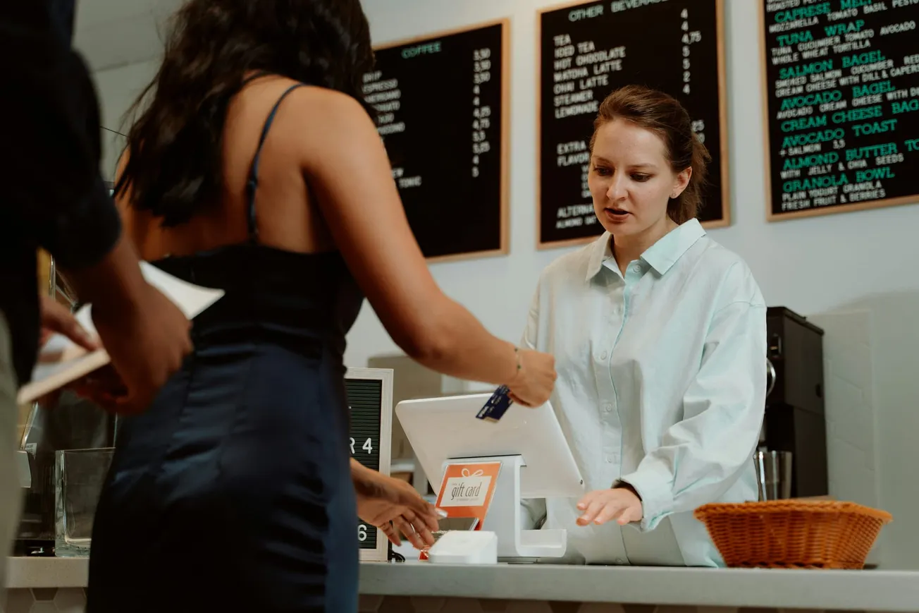 A woman in a blue dress pays with a credit card at a cafe counter. A barista in a light shirt assists her. Menu boards are visible in the background.
